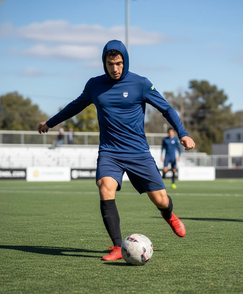 Soccer player in blue LIV.U Launch.KIT Performance long-sleeve hoodie dribbling on a sunny field.